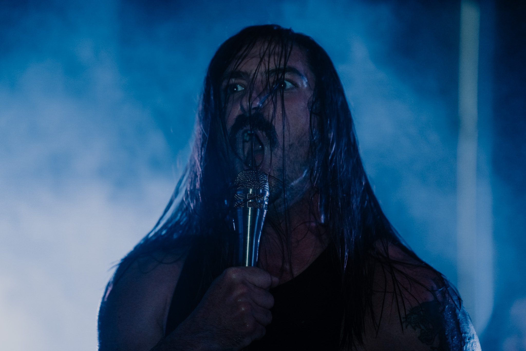 A male artist with long curly hair, wearing a black outfit, sits thoughtfully on a velvet couch with a colorful patterned background.